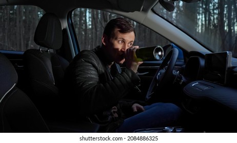 Short-haired driver sits at car, steering wheel and drinks alcohol from bottle secretly looking around for police not to see parked auto by forest