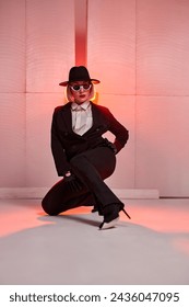short-haired modern dancer posing while sitting in studio with red backlit background. young caucasian female in black suirt, sunglasses, hat alone. portrait. grace, confidence, people, dance