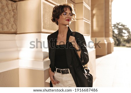 Short-haired woman in black jacket and white pants with belt posing outside. Curly girl in eyeglasses smiling at street..
