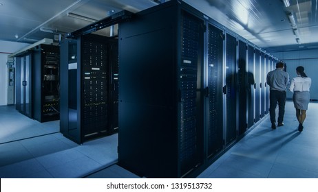 Shot of an IT Admin with a Laptop Computer and Young Technician Colleague Walking Next to Server Racks in Data Center. Running Diagnostics or Maintenance.