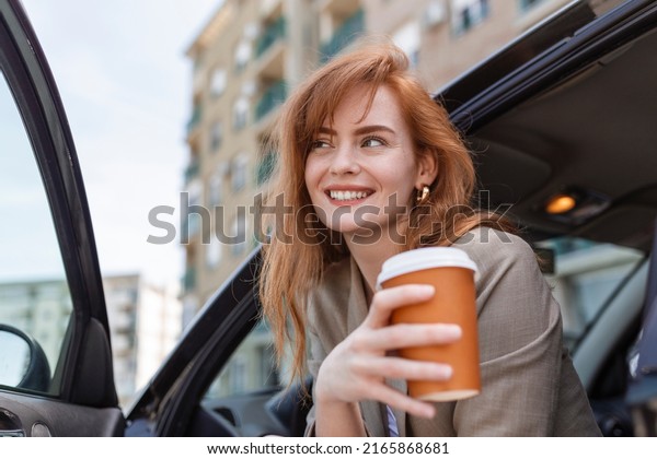 Shot of adult woman\
sitting in her car with open door, contemplating and sipping coffee\
from a travel mug.
