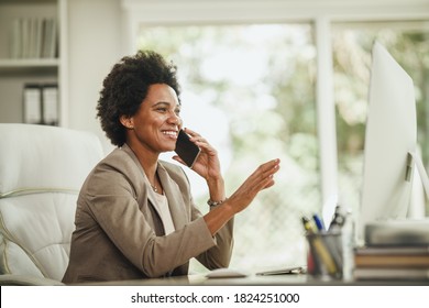 Shot of an African businesswoman talking on smartphone while working at computer in her home office during COVID-19 pandemic.