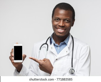 Shot Of African Doctor Isolated On Grey Background, Presenting Smart Phone And Pointing With Finger At Blank Black Screen With Copyspace For Ads