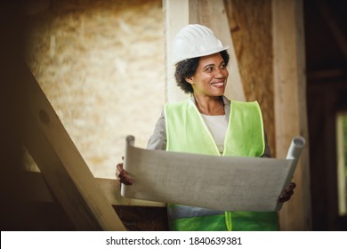 Shot Of An African Female Architect Checking Plans At The Construction Site Of A New Wooden House. She Is Wearing Protective Workwear And White Helmet.