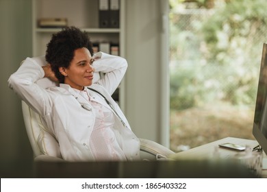 Shot Of An African Female Doctor Sitting Alone At The Desk And Relaxing After Looking X-ray On Computer In Her Consulting Room During COVID-19 Pandemic.