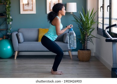 Shot of afro beautiful gymnast woman doing exercices lifting eight liters water bottles in living room at home. - Powered by Shutterstock