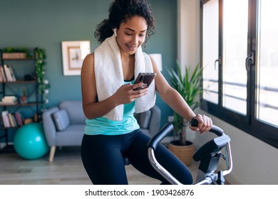 Shot Of Afro Young Fitness Girl Using Mobile Phone While Training On Exercise Bike At Home.