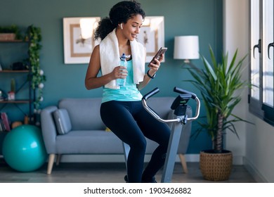 Shot of afro young fitness girl using mobile phone while training on exercise bike at home.
