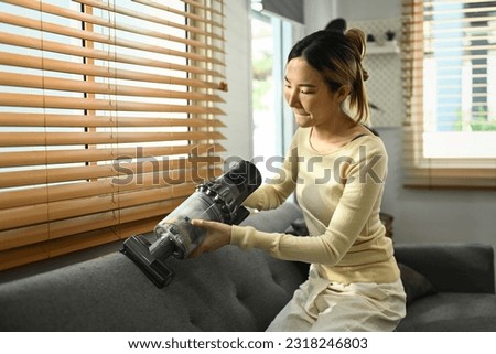 Shot of asian female housewife vacuuming sofa with cordless handheld vacuum cleaner. Housework concept