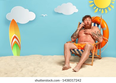 Shot of astonished bearded guy has red sunburned skin, talks on smartphone, hears shocking news from interlocutor, enjoys recreation time, sits at beach, has good rest near sea. Summer travel concept - Shutterstock ID 1438317242