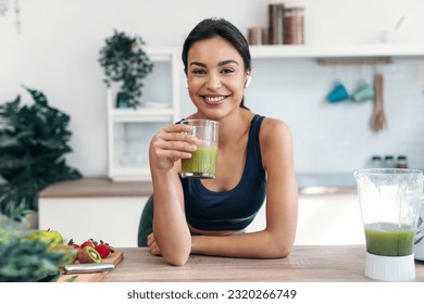 Shot of athletic smiling woman drinking smothie while listening music with earphones in the kitchen at home.
