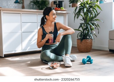 Shot of athletic woman drinking healthy protein shake while sitting on floor in the kitchen at home - Powered by Shutterstock