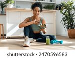 Shot of athletic woman eating a healthy fruit bowl while sitting on floor in the kitchen at home