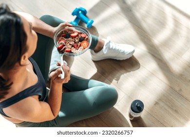 Shot of athletic woman eating a healthy bowl of muesli with fruit sitting on floor in the kitchen at home - Powered by Shutterstock