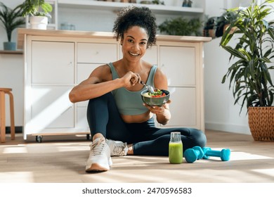 Shot of athletic woman eating a healthy fruit bowl while sitting on floor in the kitchen at home