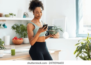 Shot of athletic woman eating a healthy fruit bowl  while using smartphone in the kitchen at home - Powered by Shutterstock