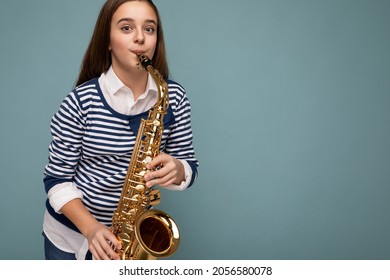 Shot of attractive positive happy brunet little girl wearing stylish striped longsleeve standing isolated over blue background wall playing saxophone looking at camera