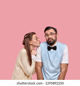 Shot Of Attractive Woman In Old Style Clothes, Gives Kiss To Clumsy Boyfriend Expresses Love And Care Pose Together Against Pink Background. Bearded Man Feels Awkward During First Date With Girlfriend