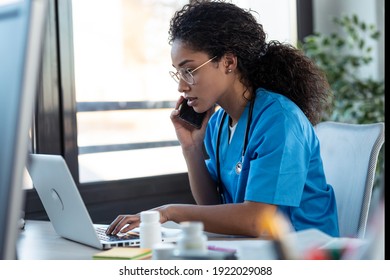 Shot of attractive young afro female doctor talking with smart phone while working with her computer in the consultation. - Powered by Shutterstock