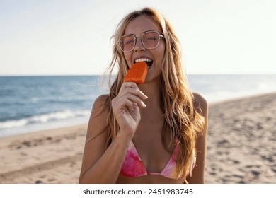 Shot of attractive young woman in bikini eating a orange popsicle looking at the camera on summer - Powered by Shutterstock