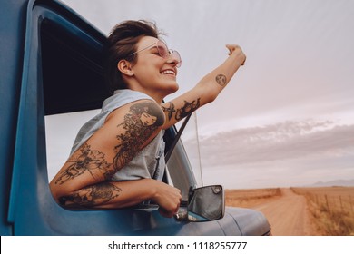 Shot Of Attractive Young Woman Enjoying Road Trip On A Summer Day. Happy Young Female Raising Her Hand Out Of The Car Window.