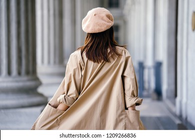 A shot of the back of a stylish Chinese Asian woman in an elegant French-inspired outfit (with a khaki trench coat and beret) in the corridor of a trendy building during the day.