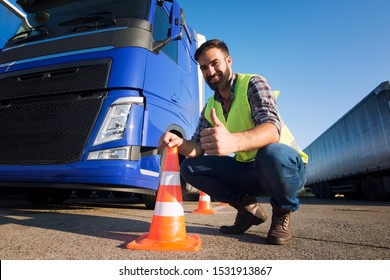 Shot Of Bearded Man Learning How To Drive Truck At Driving School. Truck Driver Candidate Training For Driving License. Standing By The Traffic Cone In Reflective Vest. Truck In The Background.