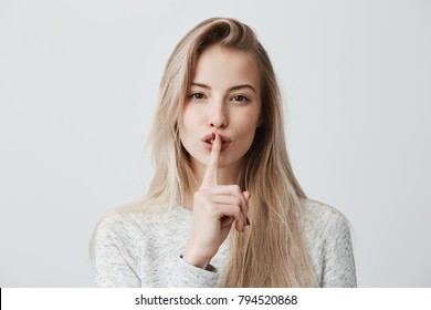 Shot of beautiful female with long blonde dyed hair wears casual long-sleeved t-shirt, shows hush sign, has serious expression, asks not to tell her secret anybody, hopes for loyality and silence.