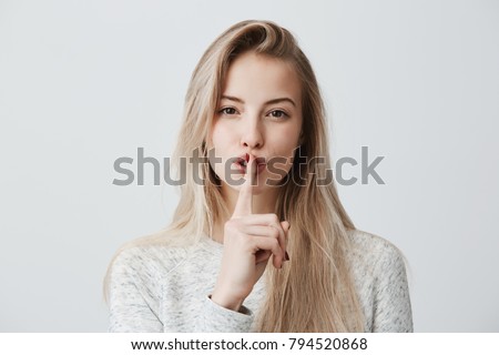 Shot of beautiful female with long blonde dyed hair wears casual long-sleeved t-shirt, shows hush sign, has serious expression, asks not to tell her secret anybody, hopes for loyality and silence.