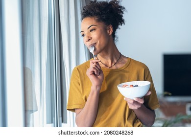 Shot of beautiful mature woman eating cereals and fruits while standing next to the window at home.