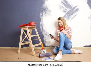 Shot Of A Beautiful Mature Woman Using Her Cellphone And Relaxing While Painting Wall In Her New Home.