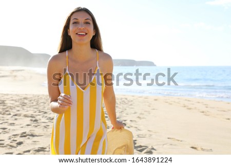 Shot of a beautiful smiling woman walking on sand beach in summer dress with hat in her hand