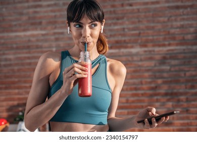 Shot of beautiful sporty woman driking a healthy smoothie while using smartphone in the kitchen.