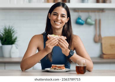 Shot of beautiful sporty woman eating healthy sanwich while looking at camera in the kitchen at home - Powered by Shutterstock