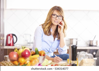 Shot of a beautiful woman making call and writing recipe into her recipe book while cooking in her kitchen.