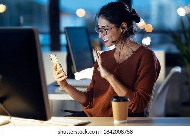 Shot Of Beautiful Young Business Woman Having Videocall With Mobile Phone While Working With Computer Sitting In The Office.