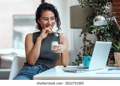 Shot of beautiful young business woman working with laptop while eating yogurt in living room at home.