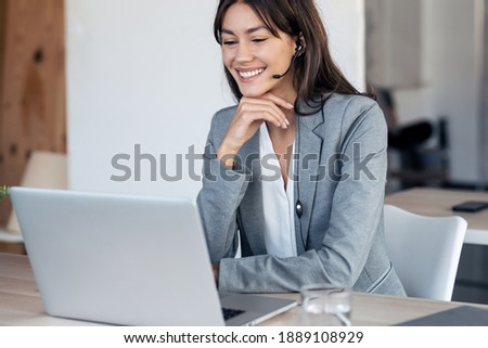 Shot of beautiful young business woman making video call with computer while talking with earphone sitting in modern startup office.