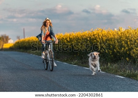 Shot of beautiful young girl riding a bike while walking her dog in the park through rapessed field.