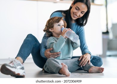 Shot Of Beautiful Young Mother Looking To Her Son While He Drinking Water From The Plastic Bottle Sitting On The Floor At Home.