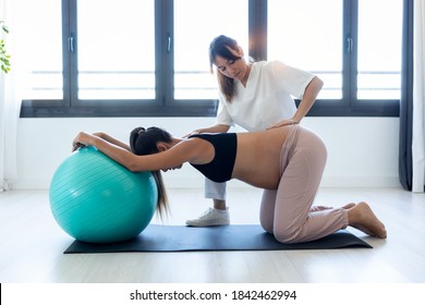 Shot of beautiful young physiotherapist helping to beautiful pregnant woman for doing pilates exercises with ball preparing for childbirth at home.