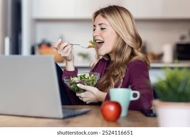 Shot of beautiful young woman doing a videocall with laptop while eating salad in the kitchen at home. - Powered by Shutterstock