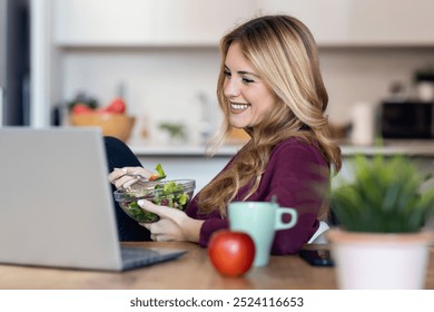 Shot of beautiful young woman doing a videocall with laptop while eating salad in the kitchen at home. - Powered by Shutterstock