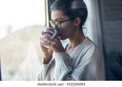 Shot of beautiful young woman drinking a cup of coffee while looking forward standing next to the window at home. - Powered by Shutterstock