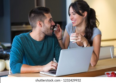 Shot of beautiful young woman giving yogurt to her handsome boyfriend while they surfing in internet with laptop in the kitchen at home. - Powered by Shutterstock