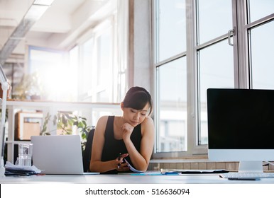 Shot Of Beautiful Young Woman Sitting At Her Work Desk Going Through Some Documents. Asian Businesswoman Working In Modern Office.