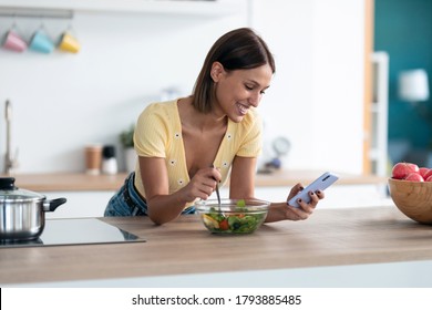 Shot of beautiful young woman using her mobile phone while eating a salad in the kitchen at home. 