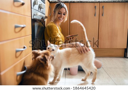 Shot of beautiful young woman using her smart phone while stroking her cute lovely cat sitting on the floor in the kitchen at home.