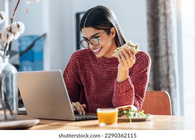 Shot of beautiful young woman working with laptop while having healthy sandwich for breakfast at home