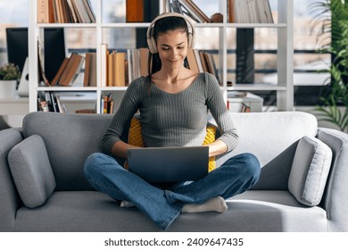 Shot of beauty young woman working with her laptop while listening to music with earphones sitting on a couch at home. - Powered by Shutterstock